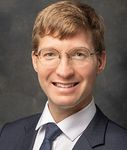 Kerry is picture from the shoulders up wearing a black collared shirt, a blue tie and eye glasses. He has fair skin, blonde hair and is smiling. 