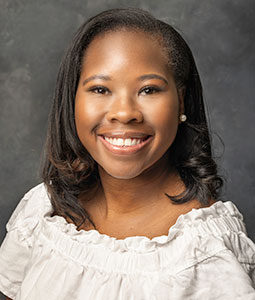 Caitlyn is pictured from the shoulders up. She has brown skin, short black hair and is wearing a white shirt. She is smiling at the camera. 