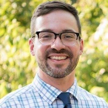 Jay is picture from the shoulders up in front of a green background. He is wearing a blue collared shirt with a blue tie, eyeglasses, and is smiling. 