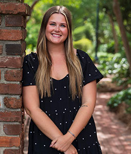 Ryanne is pictured from the waist up wearing a black and white polka dot dress. She has long blonde hair, fair skin and is smiling. 