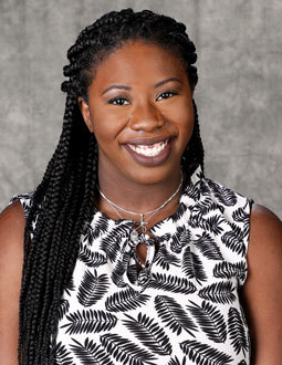 Kelsey is pictured from the shoulders up wearing a black and white floral shirt. She has long braided hair and she is smiling. 