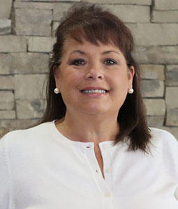 Marilee is pictured from the shoulders up wearing a white shirt. She has short brown hair, brown eyes, pale skin and is smiling. 