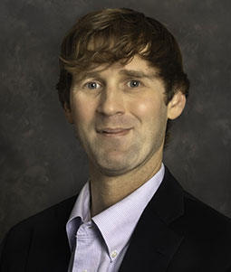 Neal is pictured from the shoulders up wearing a white collared shirt and black jacket. He has blonde-brown hair and brown eyes. 