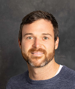 Charles is pictured from the shoulders up wearing a blue shirt. He has pale skin, brown hair and a beard. He is smiling. 