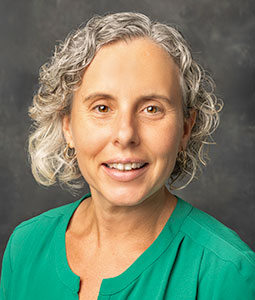 Kim is pictured from the shoulders up wearing a green shirt. She has tan skin, gray curly hair and is smiling. 