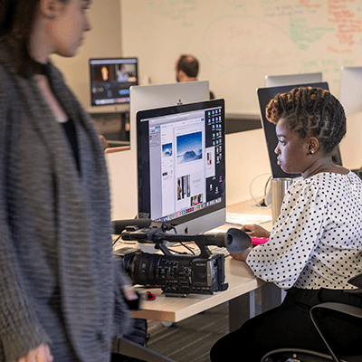 A student working at a computer in a computer lab with a large camera on the desk next to her.