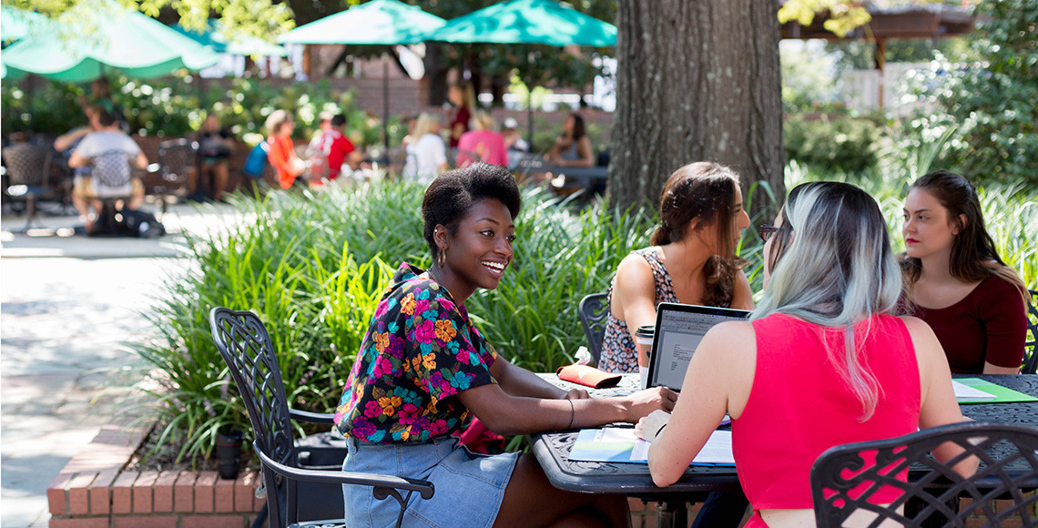 Students sit in the shade outside Russell House with an open laptop.