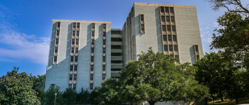 A front view of Bates House, a building with blue/grey bricks and windows lining the building in columns, with green trees in the foreground.