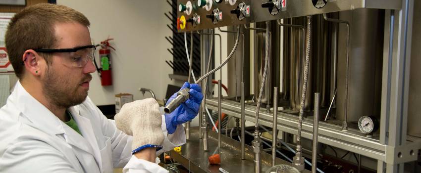 Male student in white lab coat and safety glasses works on lab equipment