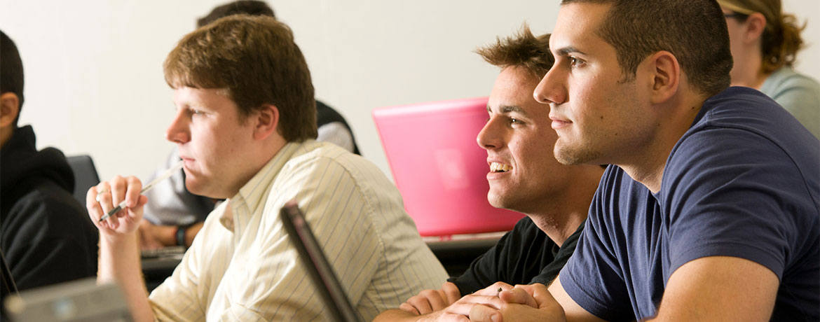 Close-up of six students in a classroom, leaning forward at their desks, listening intently to a lecture