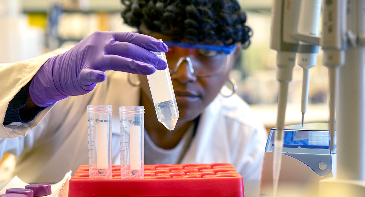 A student wearing safety goggles, gloves and a lab coat examines a test tube full of clear liquid. 