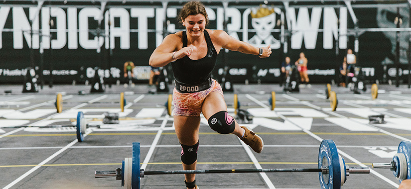 Reese Littlewood, wearing a black tank top and patterned shorts, runs toward the finish line in a CrossFit competition, surrounded by weightlifting equipment on a gym floor with 'Syndicate Crown' branding in the background.