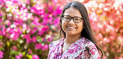 A woman with glasses and long hair smiling in front of vibrant pink flowers.