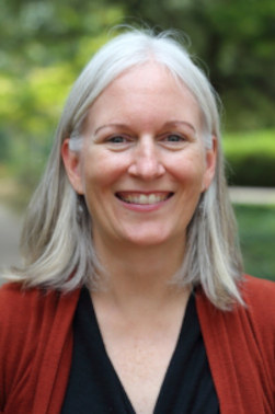 Kate Flory is smiling while wearing a black top and an orange cardigan. She is posed in front of a blurred green background. 