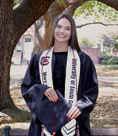 Marygail standing in front of tree on the Horseshoe in graduation regalia.