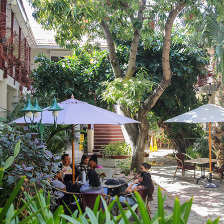 Students at the University of Aruba site at a table under an umbrella in a common space surrounded by lush vegetation.