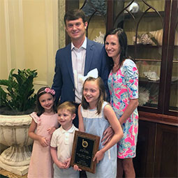 Blake Taylor stands with his family inside their home. His older daughter holds his award.