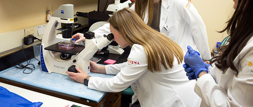 Female medical student in white coat smiling in lecture hall. One deliberately different medical school logo.