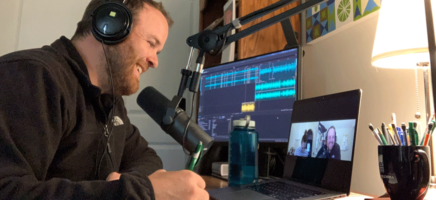 Visual communications instructor Jason Porter wears headphones and sits in front of a microphone and laptop