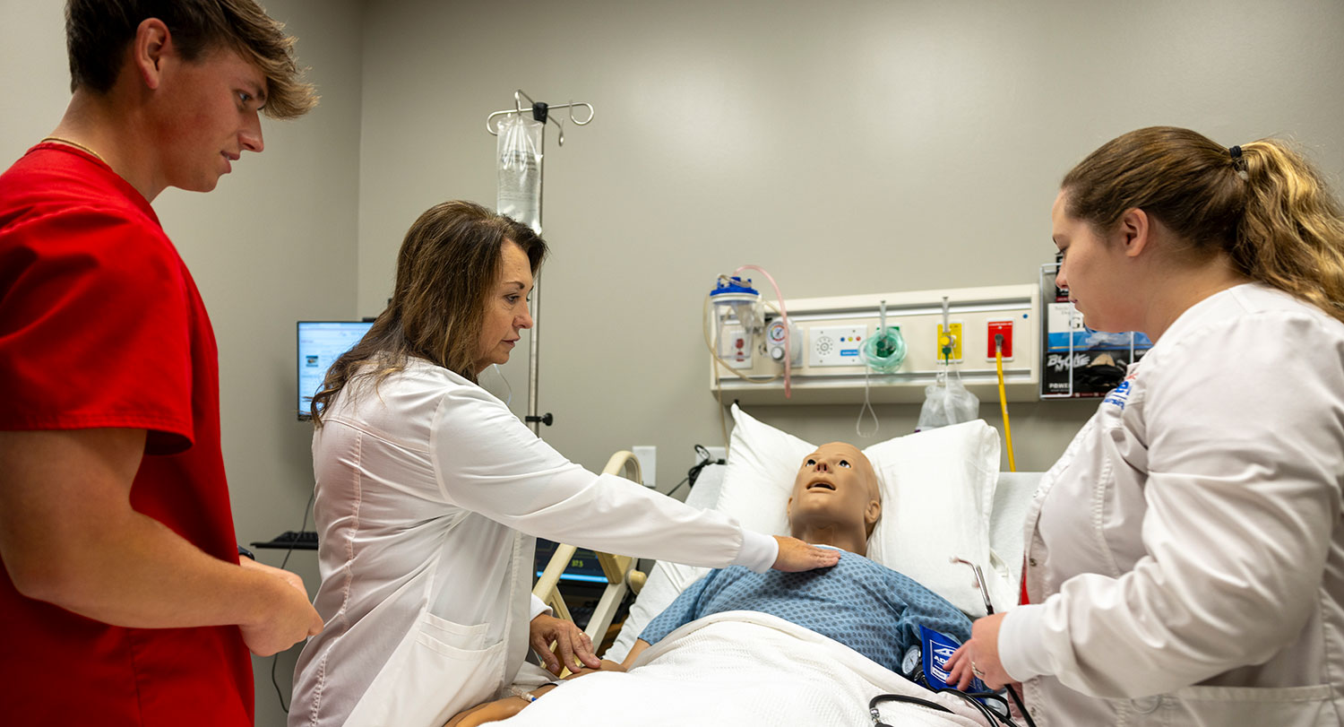 An instructor stands over a high-fidelity mannequin while two students stand nearby