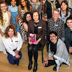 Author Julia Elliott, brown shoulder-length hair and wearing a brown suit, sits in a chair holding her book surrounded by group of onlookers
