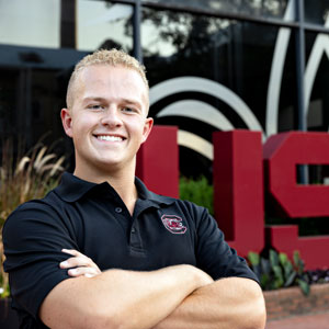 A smiling man in USC polo shirt poses with crossed arms in front of university signage.