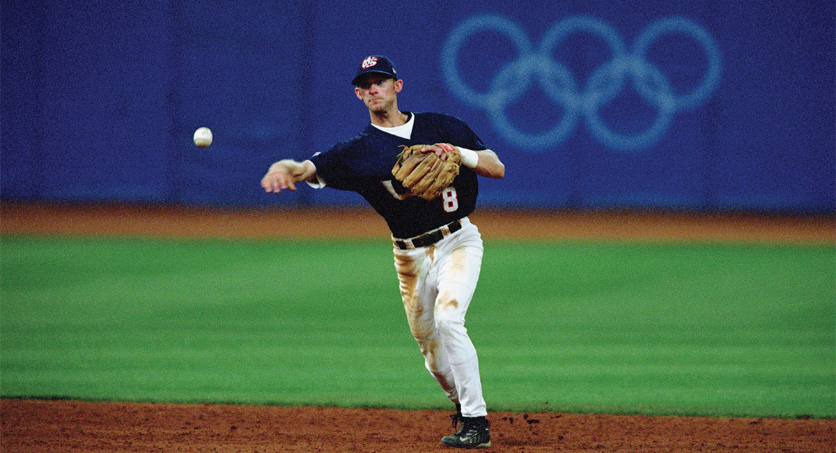 man throws a baseball on the field with the olympic rings in the background